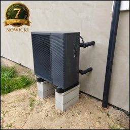 GREEN ENERGY Sp. z.o.o. Pompy Ciepła Nowicki Technika Grzewcza - Szara pompa ciepła na betonowych blokach przy ścianie budynku z widoczną rurą spustową. Na pompie naklejka z gwarancją. Teren wokół w trakcie prac ziemnych.