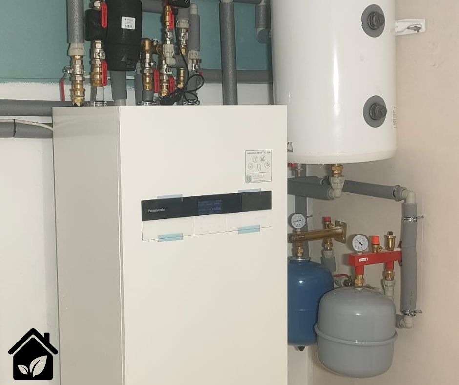Biała jednostka wewnętrzna z wyświetlaczem Panasonic Aquarea Smart Cloud, zbiornik buforowy i osprzęt instalacyjny w kotłowni.
