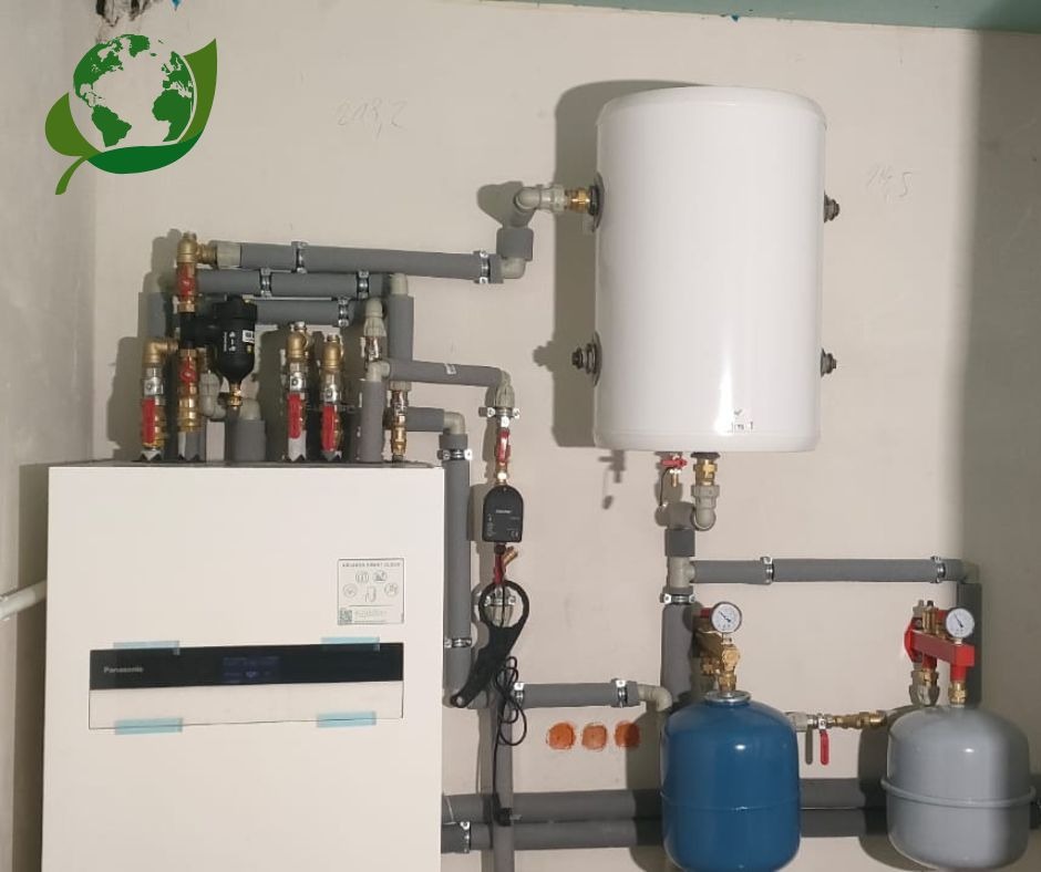 Instalacja grzewcza w kotłowni z białą jednostką Panasonic, zbiornikiem buforowym, naczyniami przeponowymi i szarymi rurami PCV, widoczne manometry i zawory, surowy stan wykończenia ścian.