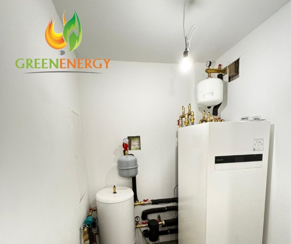 Białe pomieszczenie z zainstalowaną białą pompą ciepła Panasonic, zbiornikami wyrównawczymi i rurami, widoczne logo firmy GREENENERGY w lewym górnym rogu.