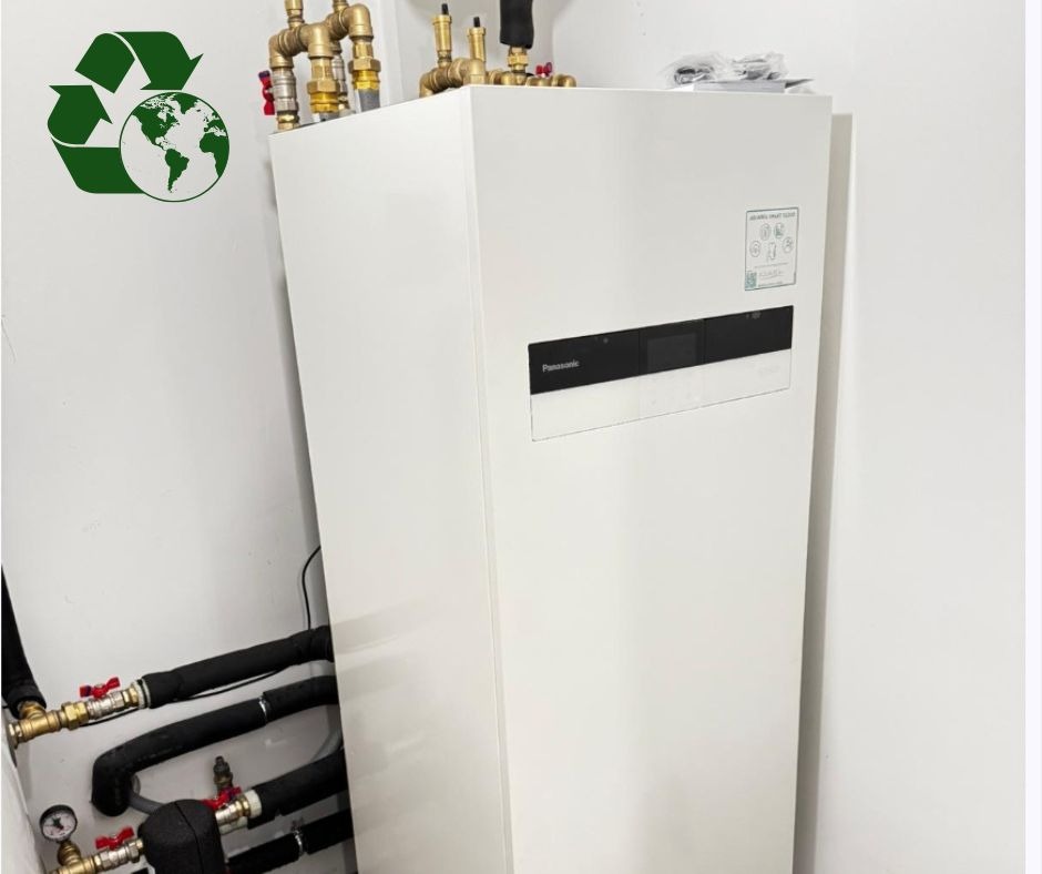 Biała jednostka wewnętrzna systemu grzewczego Panasonic, z widocznymi przyłączami hydraulicznymi z miedzianymi zaworami i czarnymi izolowanymi rurami, umieszczona w rogu pomieszczenia z białymi...