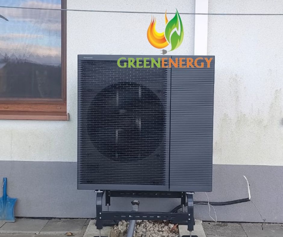 Zewnętrzna jednostka pompy ciepła Panasonic, zamontowana na metalowej konstrukcji przy ścianie budynku z widocznym oknem i logo firmy Greenenergy na górze ściany.