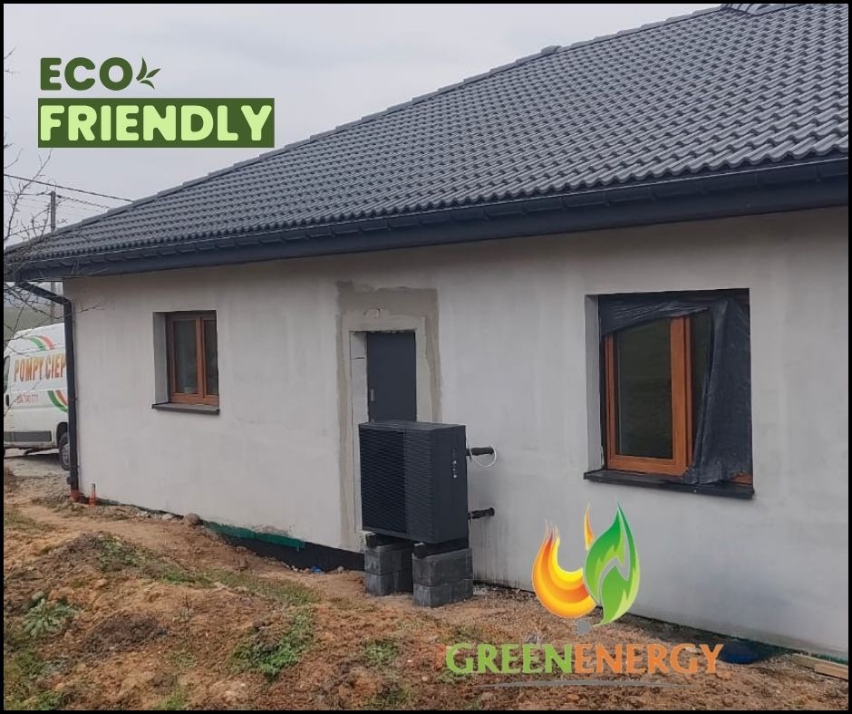 Zewnętrzna jednostka pompy ciepła zamontowana przy ścianie domu z szarą dachówką, widoczny fragment firmowego samochodu dostawczego z napisem 'Pompy Ciepła' w tle, okno zabezpieczone folią.