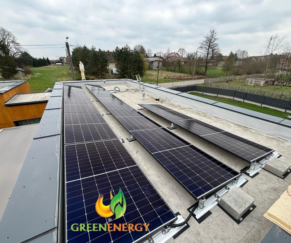 Panele fotowoltaiczne na płaskim dachu domu jednorodzinnego, zamontowane na aluminiowej konstrukcji z betonowymi obciążnikami. Widok z góry, pochmurne niebo.