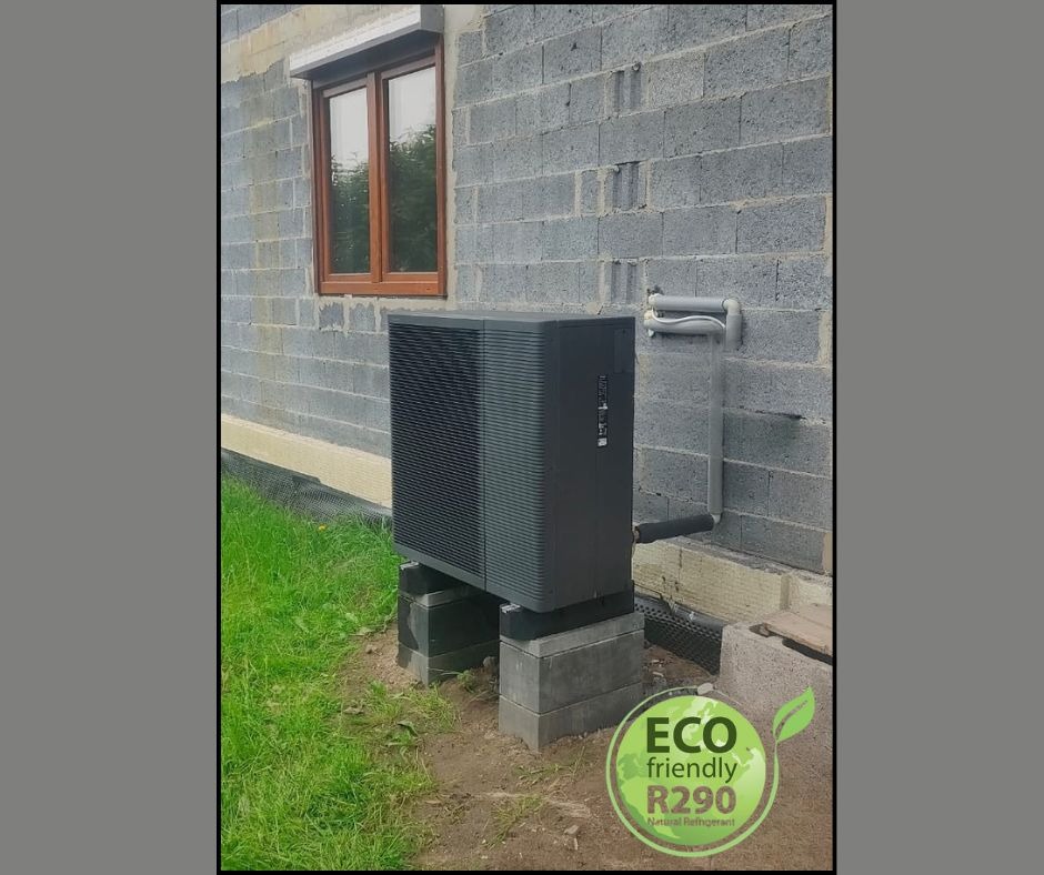 Czarna pompa ciepła na betonowych blokach przy szarym budynku z oknem. Widoczne rury i logo 'Eco friendly R290'.