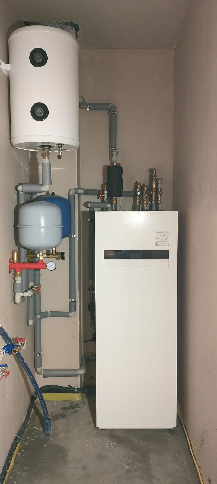 Zmontowana instalacja grzewcza z białym zbiornikiem i szarymi rurami w pomieszczeniu o beżowych ścianach. Widoczny manometr i zawory.