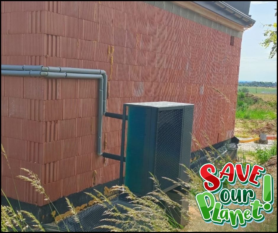 Zewnętrzna jednostka pompy ciepła zamontowana przy ceglanej ścianie budynku. Widoczne rury przyłączeniowe i fragment krajobrazu w tle. Naklejka 'Save our planet'.
