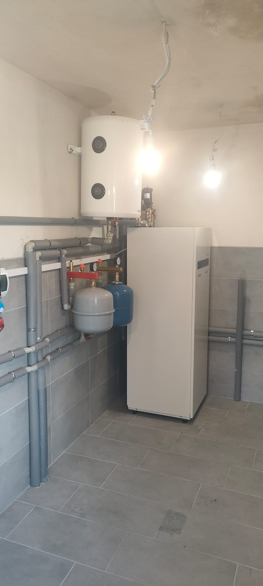 Zmontowana instalacja grzewcza w pomieszczeniu z szarymi płytkami. Widoczny zasobnik ciepłej wody, pompa ciepła i orurowanie. Oświetlenie sufitowe w trakcie montażu.