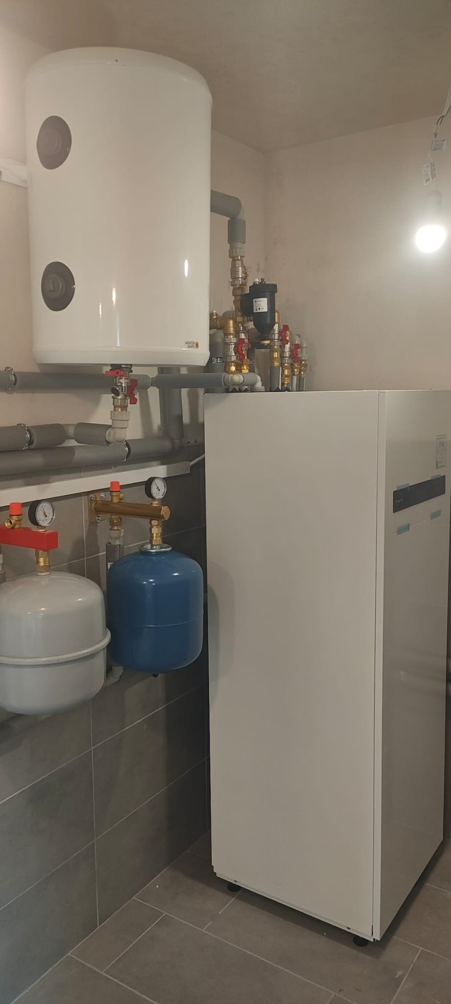 Biały zasobnik ciepłej wody i pompa ciepła w kotłowni z szarą glazurą. Widoczne rury, zawory i manometry. Oświetlenie sufitowe.
