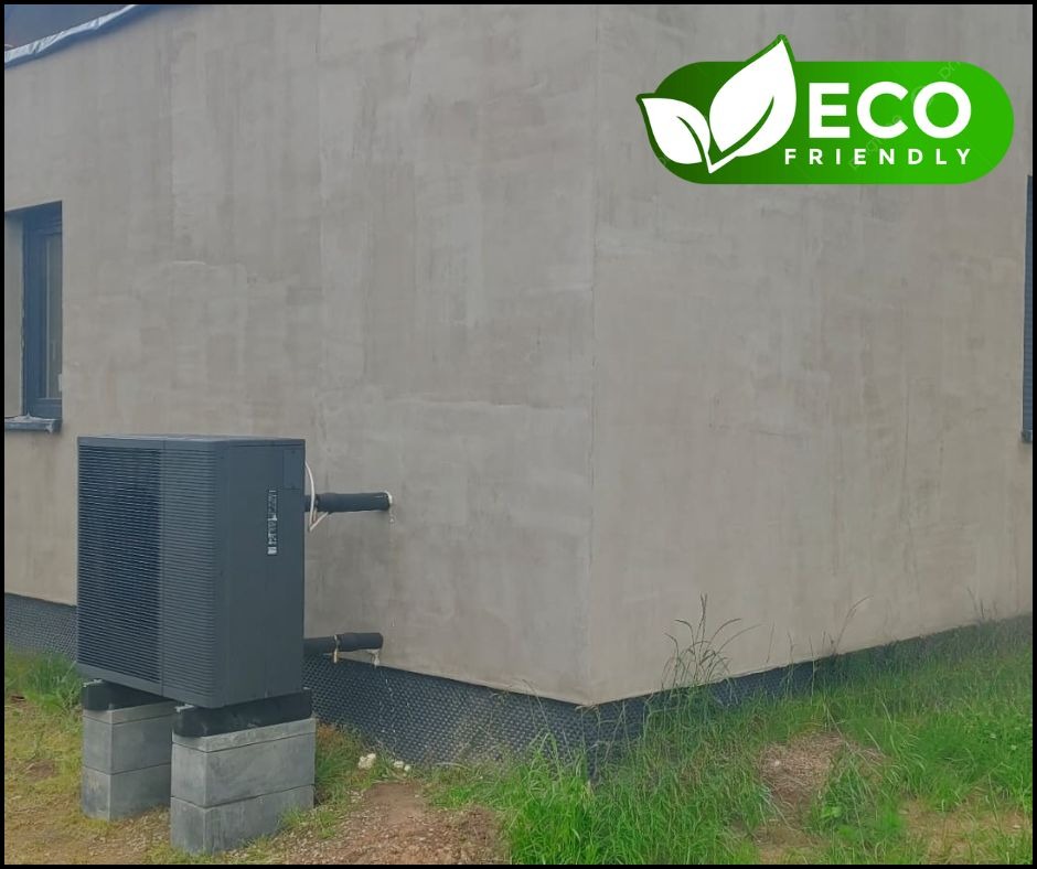 Czarna pompa ciepła na betonowych blokach przed surową elewacją budynku z logo 'Eco Friendly' w prawym górnym rogu.