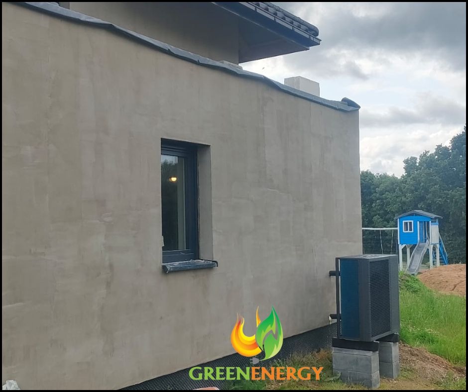 Zewnętrzna jednostka pompy ciepła zamontowana przy ścianie domu z widocznym oknem i placem zabaw w tle. Szara elewacja budynku. Logo firmy GREENENERGY w rogu.