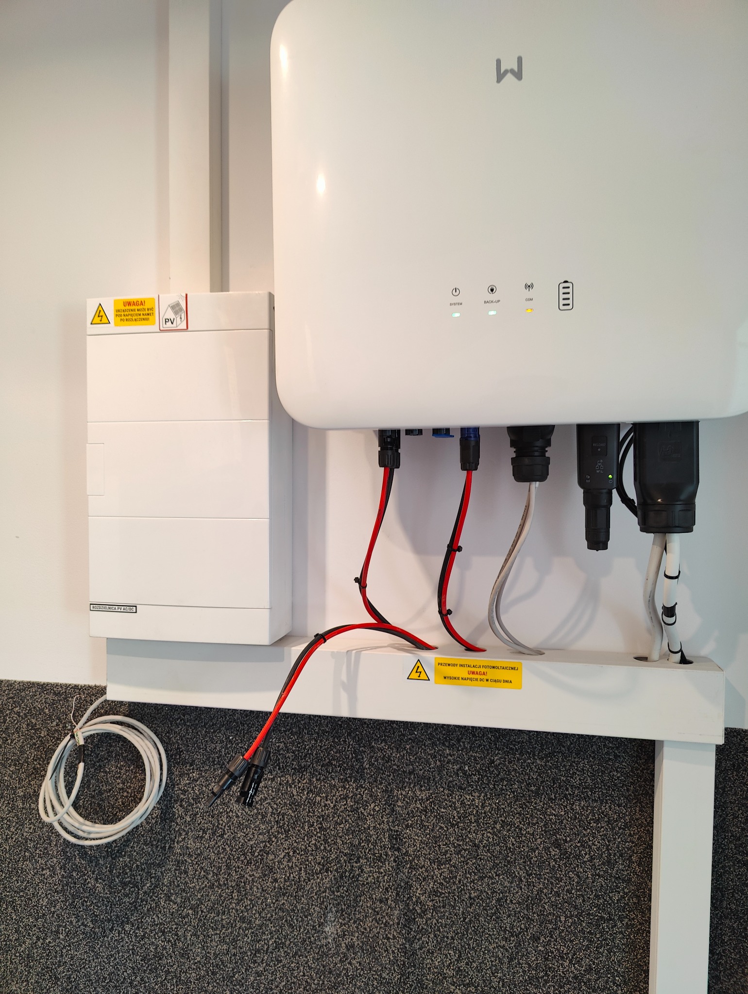 Biała skrzynka rozdzielcza PV AC/DC z żółtą naklejką ostrzegawczą. Pod spodem widoczne kable instalacji fotowoltaicznej.