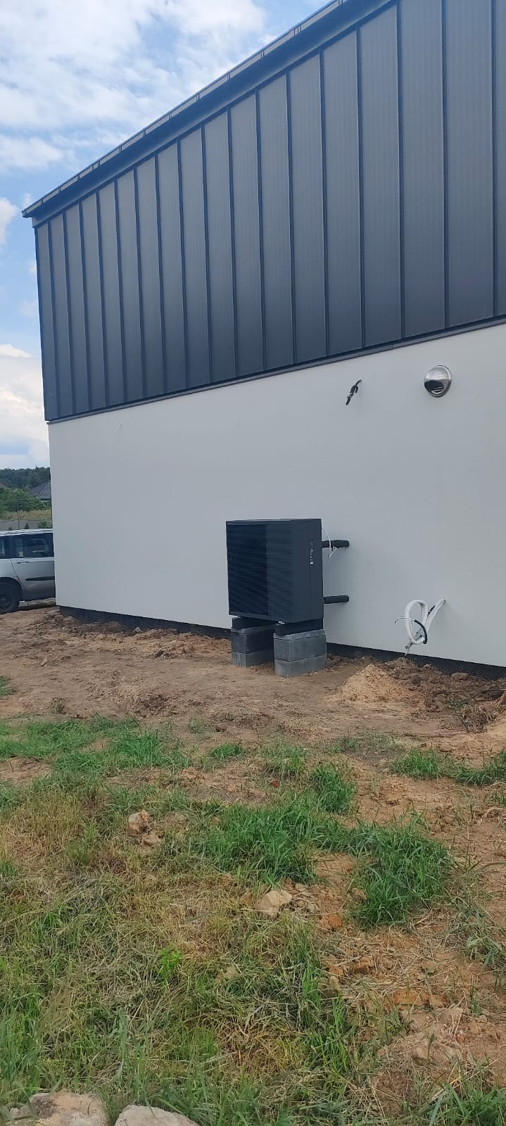 Czarna pompa ciepła zamontowana na betonowych bloczkach przy białej ścianie budynku z szarą blachą trapezową na dachu. Widoczna instalacja oraz trawa z ziemią.