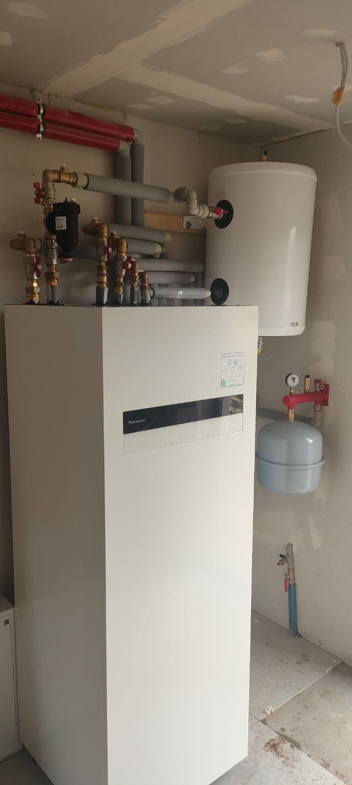 Biała pompa ciepła Panasonic z zbiornikiem buforowym i instalacją hydrauliczną w pomieszczeniu z surowym wykończeniem ścian i podłogi.