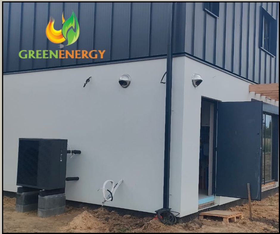Zamontowana, czarna pompa ciepła na betonowych blokach przy budynku z otwartymi, ciemnymi drzwiami. Widoczny fragment elewacji i logo 'Greenenergy'.