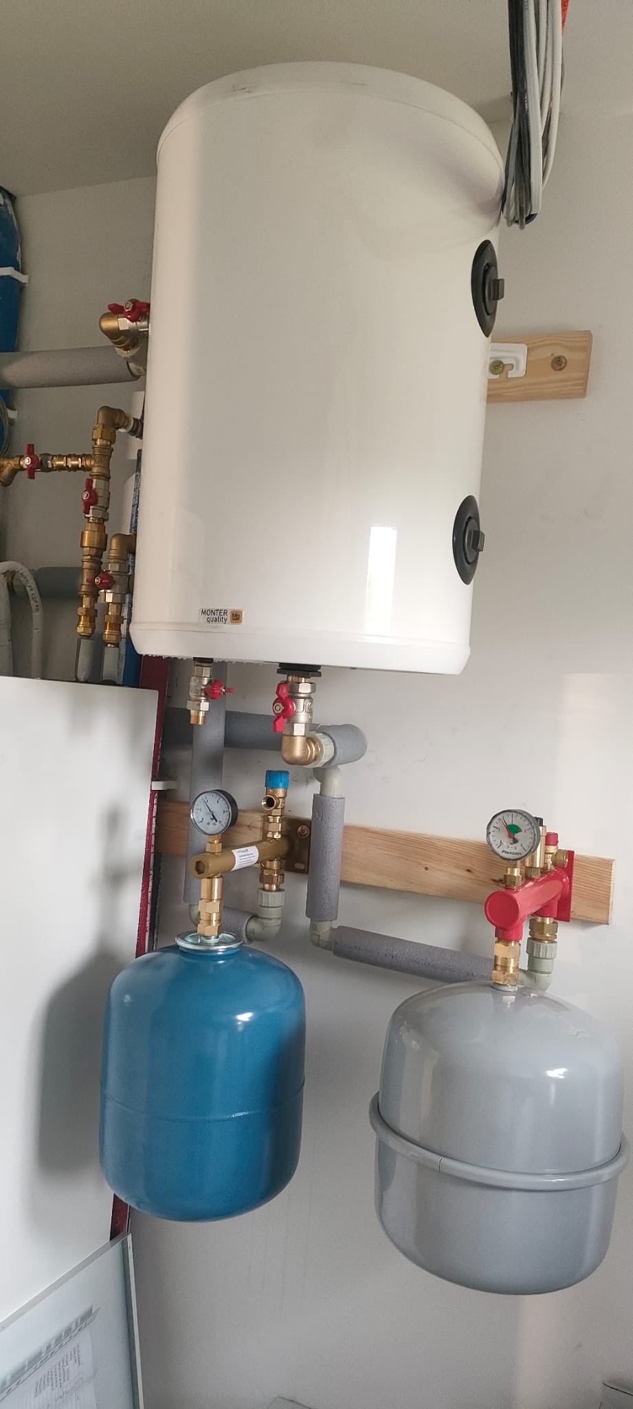 Biały zasobnik ciepłej wody użytkowej z osprzętem, manometrami i zaworami, zamontowany na ścianie, z widocznymi zbiornikami wyrównawczymi.