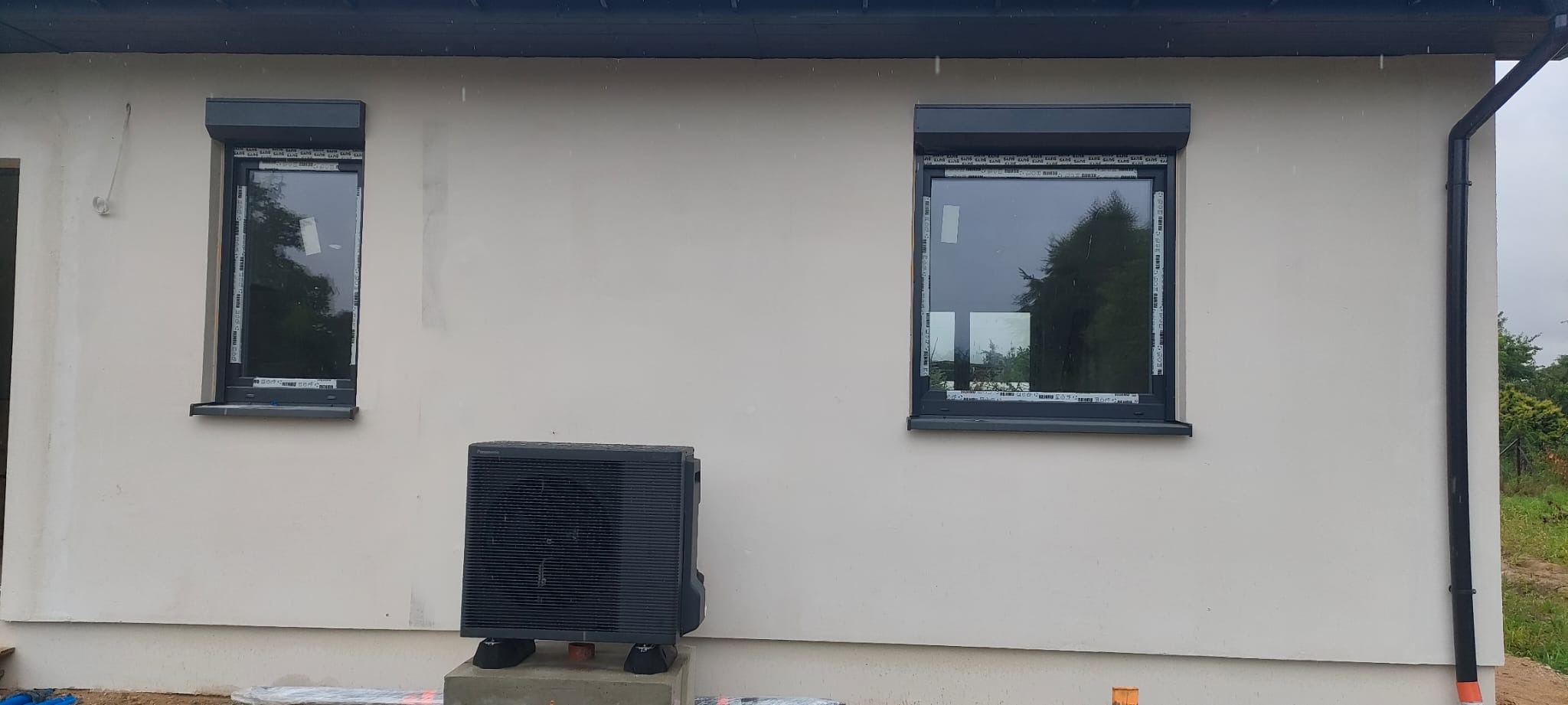 Zamontowana czarna pompa ciepła Panasonic na zewnątrz budynku z dwoma oknami z roletami. Widoczne orynnowanie. Pochmurna pogoda.