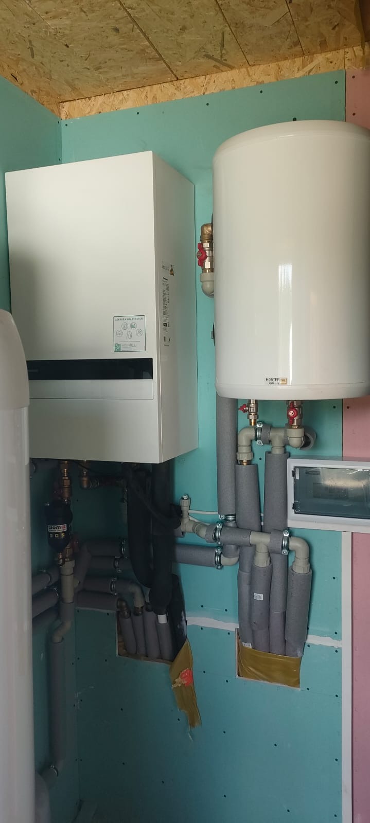 Biała pompa ciepła Aquarea i zbiornik Monterm Quality z szarymi rurami w pomieszczeniu z płytami gipsowymi. Instalacja w trakcie wykończenia.