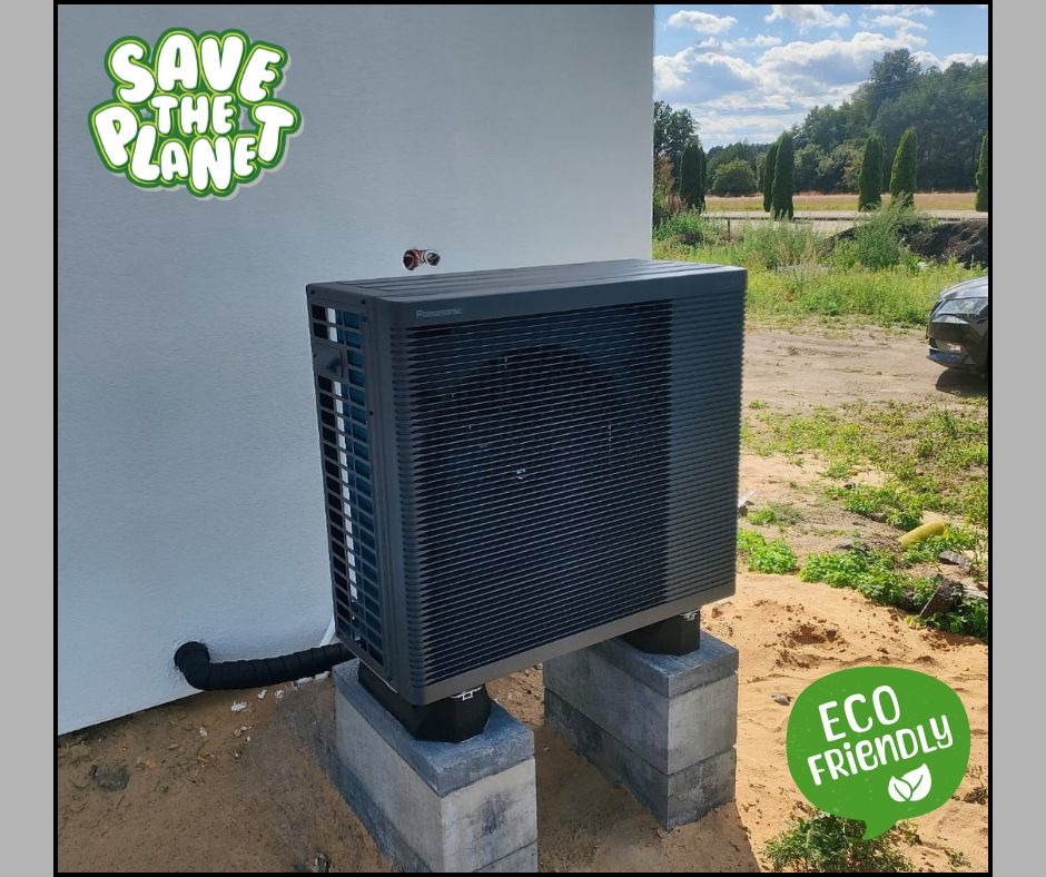 Szara pompa ciepła Panasonic, zamontowana na betonowych blokach przy białej ścianie budynku, z naklejkami 'Save the planet' i 'Eco friendly'.