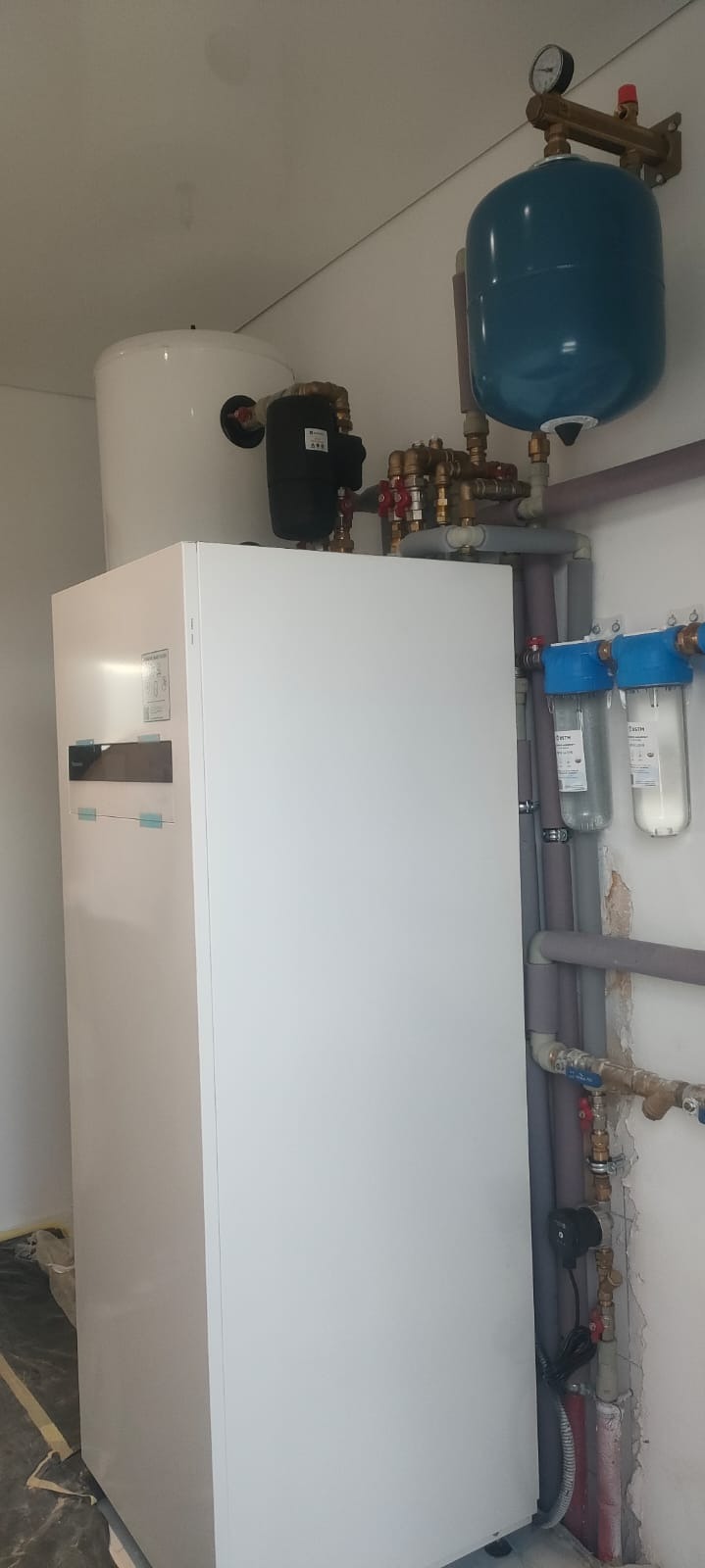 Biała pompa ciepła z zbiornikiem, zaworami i filtrami wody. Szare rury instalacji grzewczej na ścianie. Widok na kompletną instalację w pomieszczeniu.