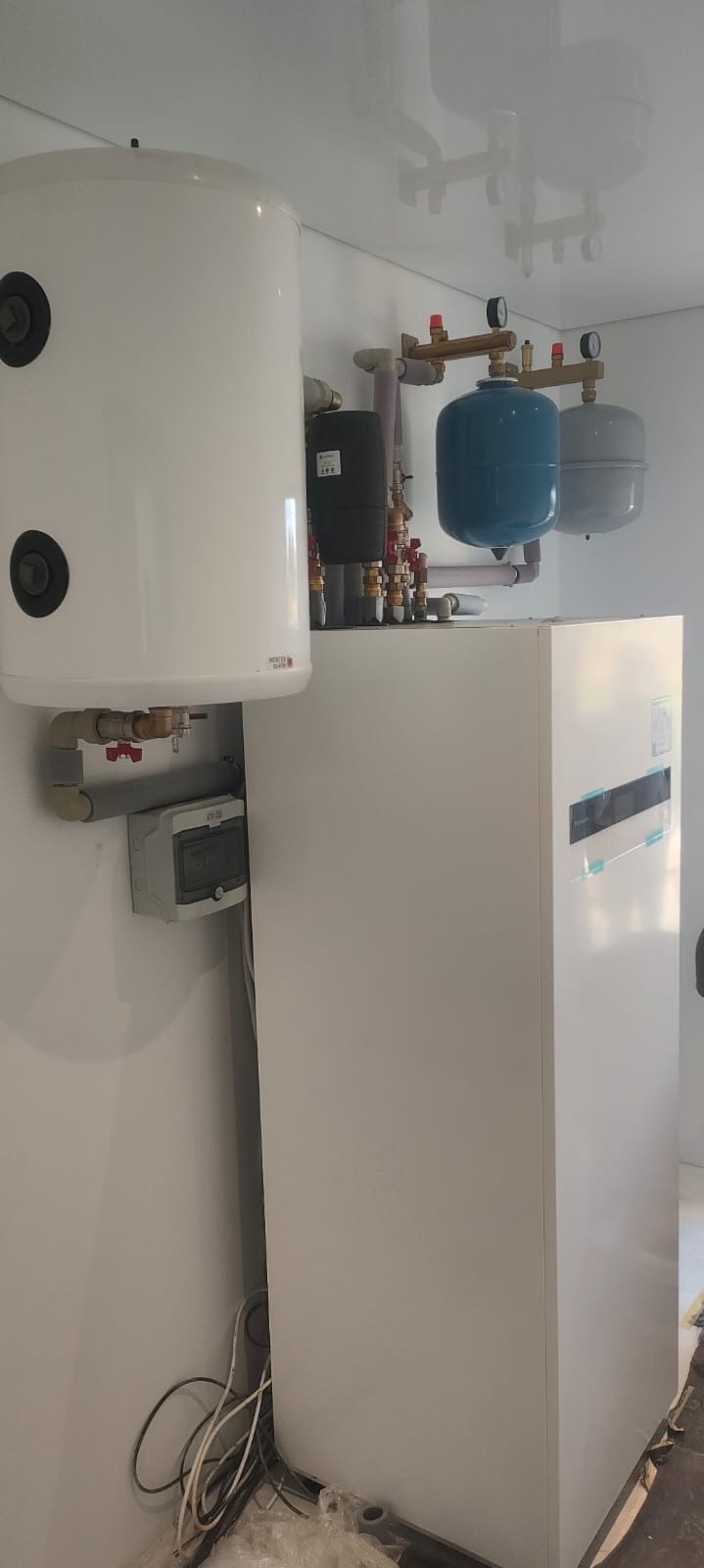 Biały zasobnik ciepłej wody użytkowej, obok biała pompa ciepła, instalacja z rur i zaworów, szare naczynia przeponowe. Całość w jasnym pomieszczeniu.