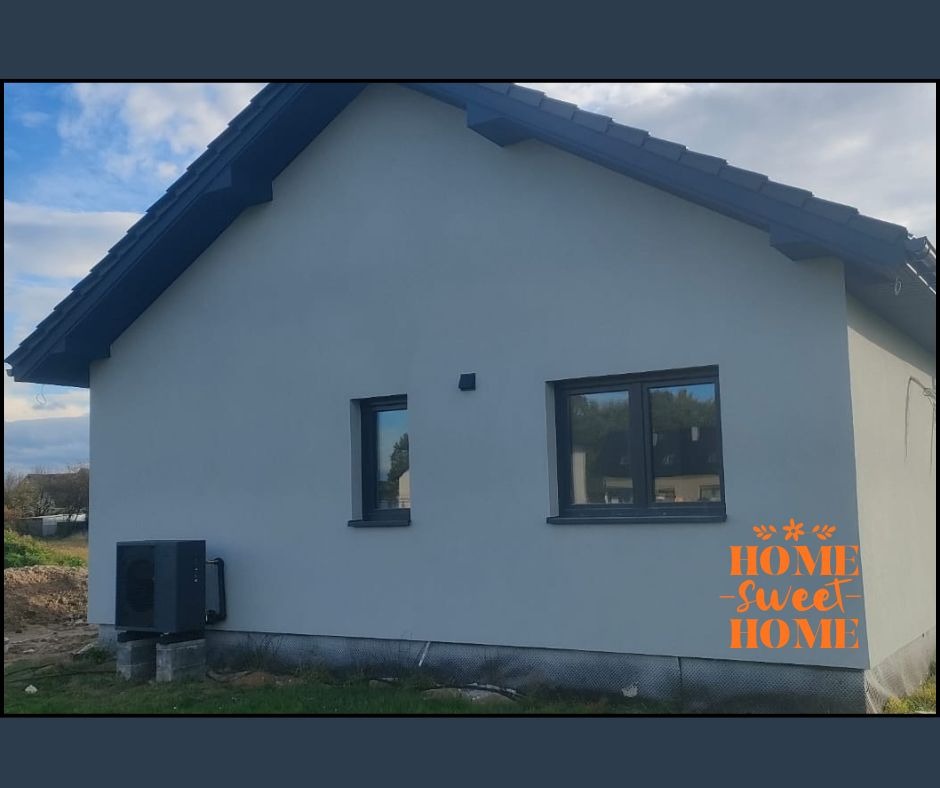 Szary dom jednorodzinny z zamontowaną czarną pompą ciepła na zewnątrz. Widoczne okna i dach. Na ścianie pomarańczowy napis 'Home Sweet Home'.