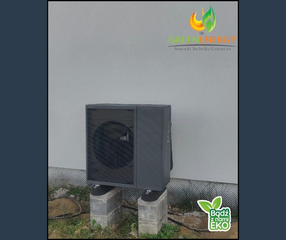 Szara pompa ciepła zamontowana na betonowych blokach przy jasnej ścianie budynku. Widoczne logo firmy Green Energy i hasło 'Bądź z nami EKO'.