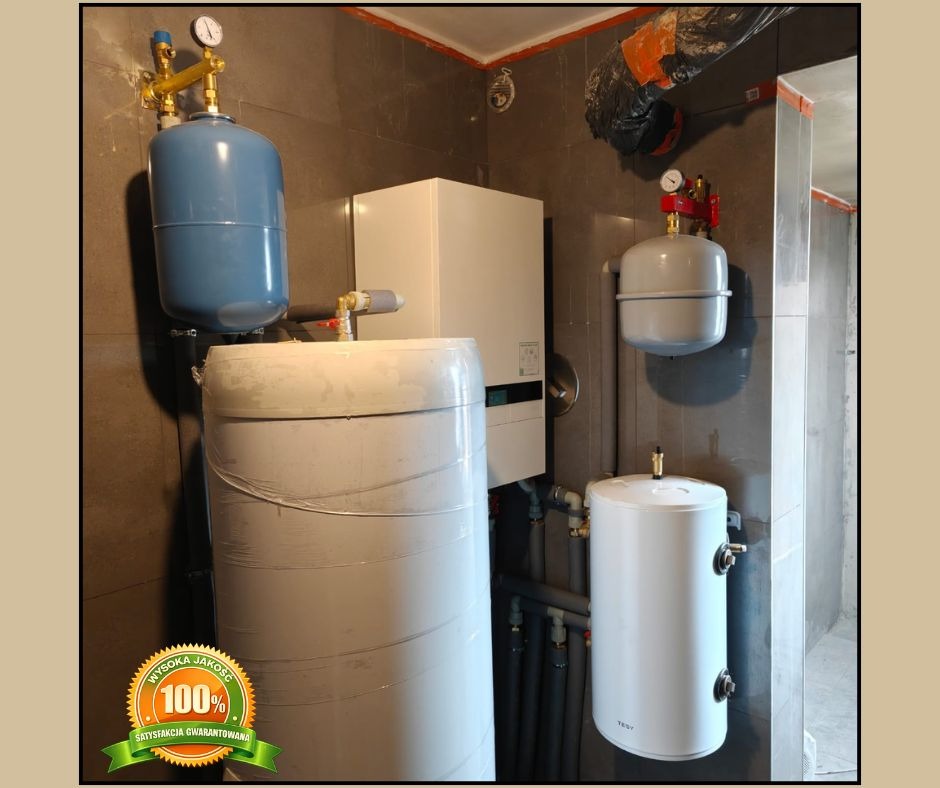 Zmontowana instalacja grzewcza w kotłowni z szarymi płytkami. Widoczne zbiorniki, rury i piec. Na jednym ze zbiorników naklejka '100% satysfakcji'.