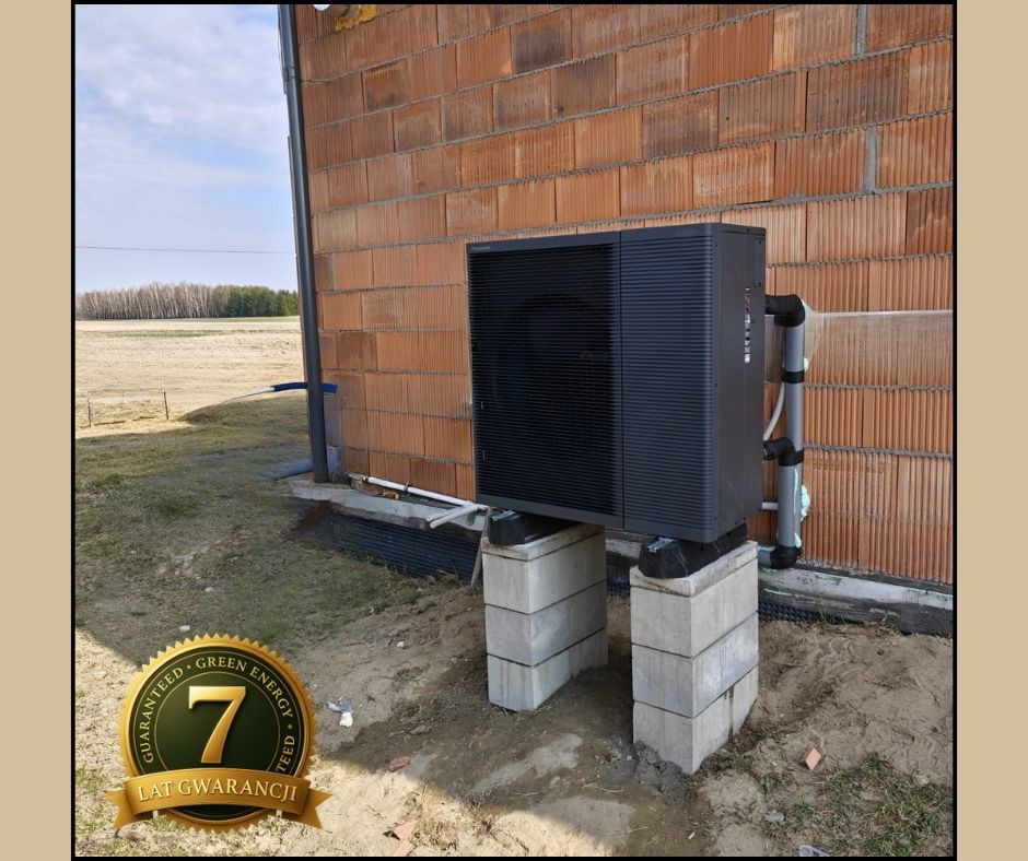 Czarna pompa ciepła powietrze-woda, zamontowana na betonowych blokach przy ceglanej ścianie budynku w trakcie montażu. Widoczny krajobraz polny w tle.