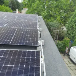 Panele słoneczne na płaskim dachu z widocznymi elementami montażowymi i fragmentem instalacji elektrycznej; w tle zieleń drzew i pracownik obok białego busa.