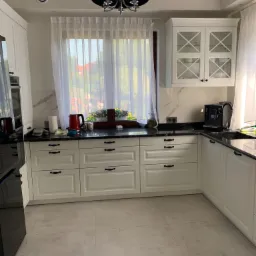 Jasna kuchnia z białymi szafkami, czarnym blatem i marmurowym wykończeniem ściany, okno z firanką i kwiatami, nowoczesna czarna lampa wisząca.