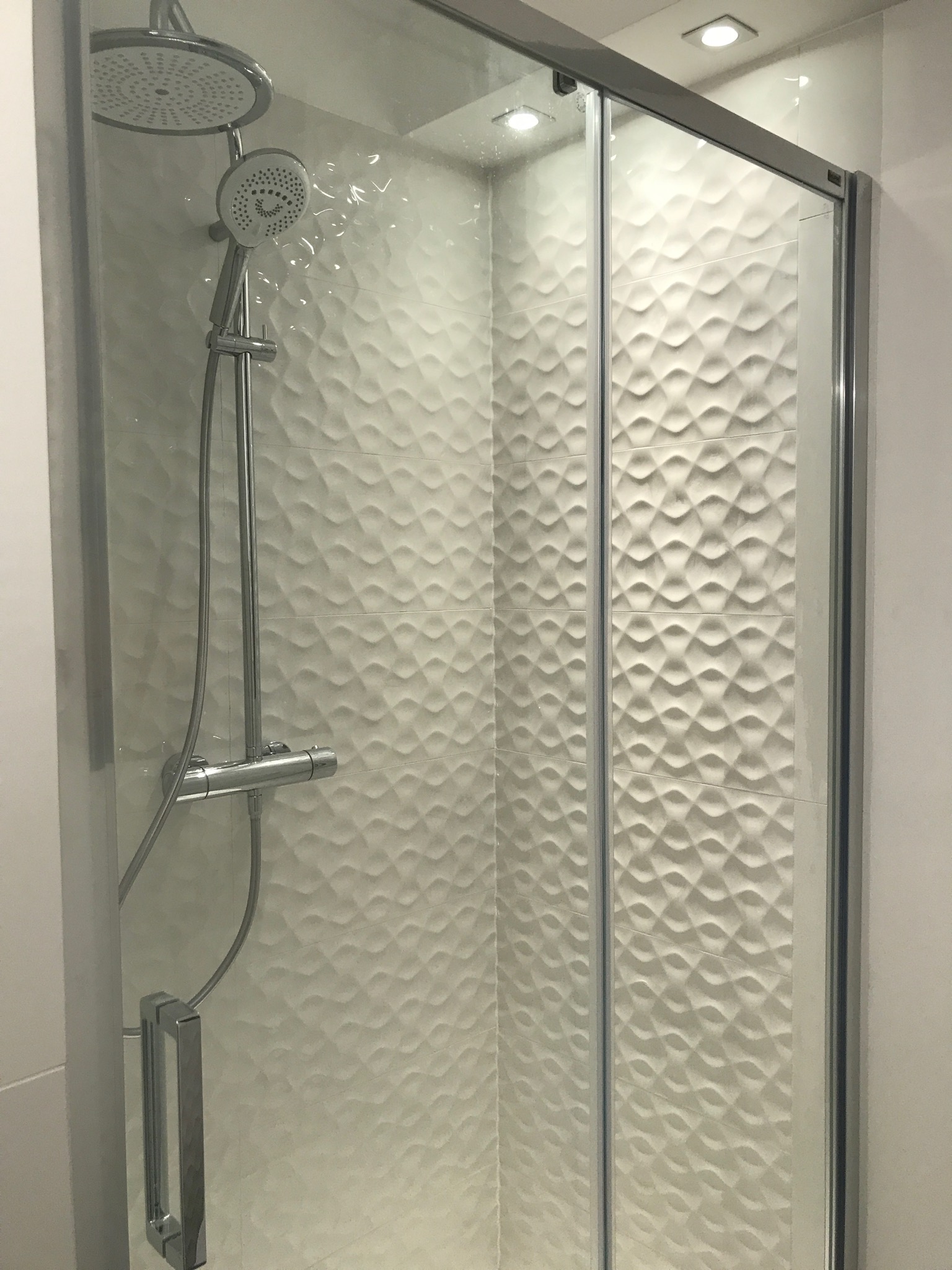 Kabina prysznicowa z deszczownicą i panelem z teksturalnych, białych płytek ceramicznych, oświetlona punktowo.