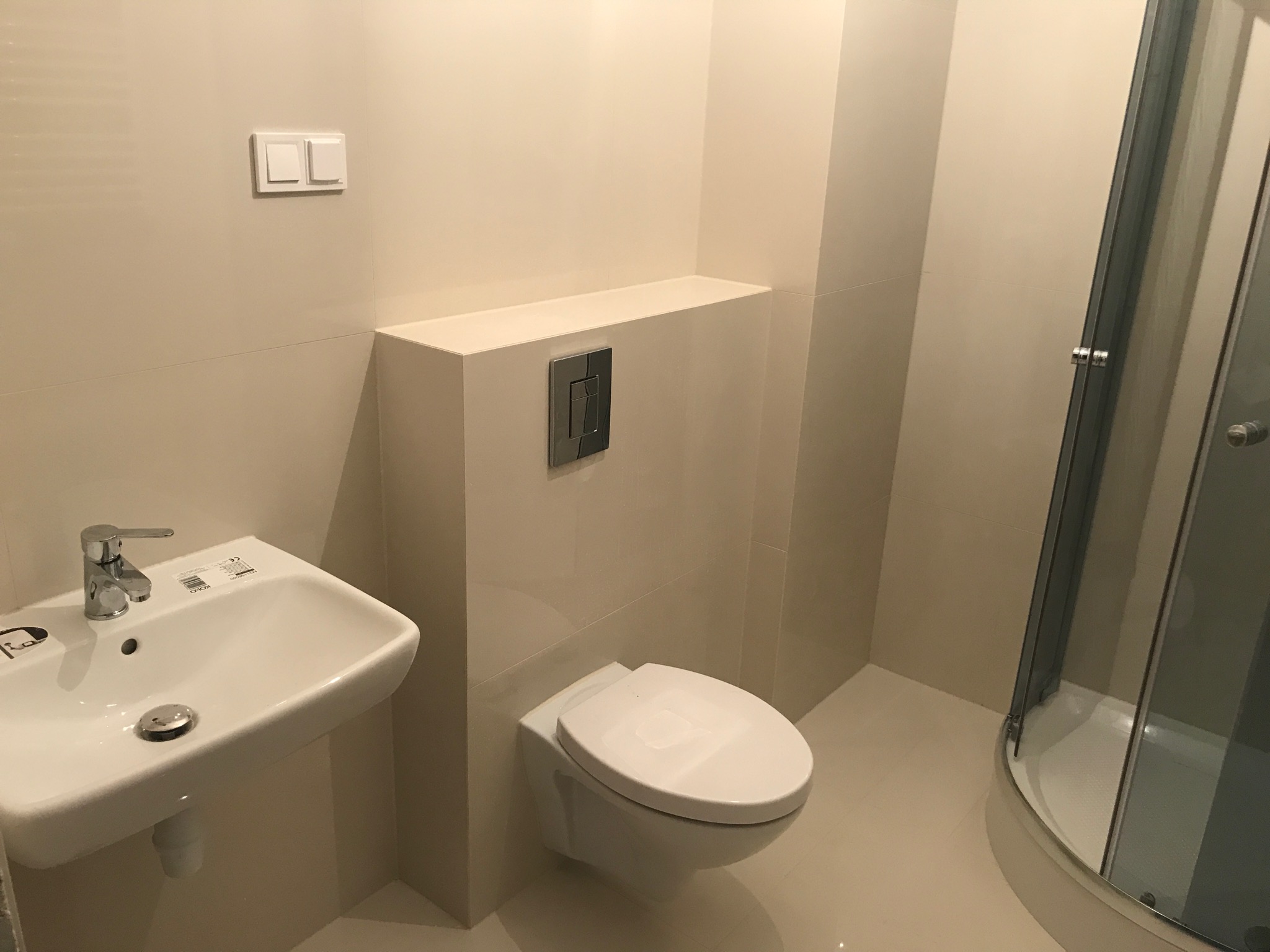 Biała, minimalistyczna łazienka z podwieszaną toaletą, umywalką Kolo i kabiną prysznicową z ciemnymi profilami, wykończona jasnymi, błyszczącymi płytkami.