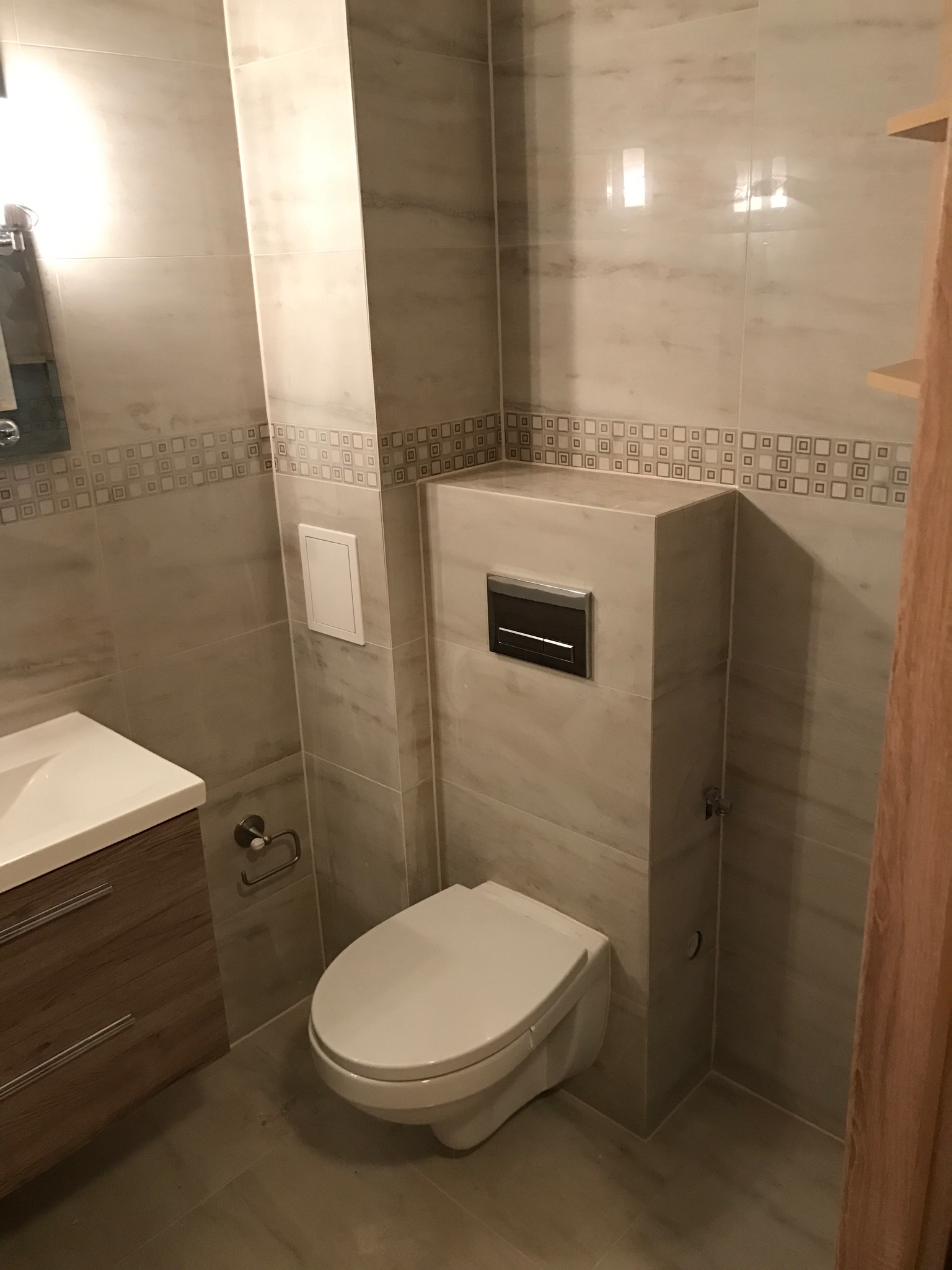 Wykończona łazienka z beżowymi płytkami imitującymi marmur, wisząca toaleta z zabudowanym systemem spłukiwania, umywalka z szafką w kolorze drewna i dekoracyjnym pasem płytek z motywem geometrycznym.