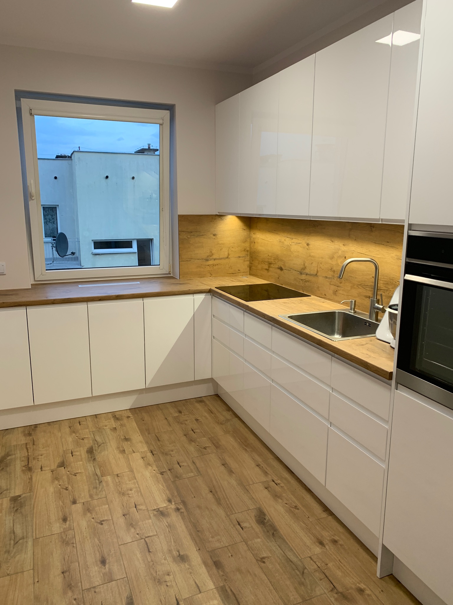 Nowoczesna kuchnia z białymi, lakierowanymi szafkami, drewnianym blatem i panelami, widok na okno z budynkiem mieszkalnym i anteną satelitarną.