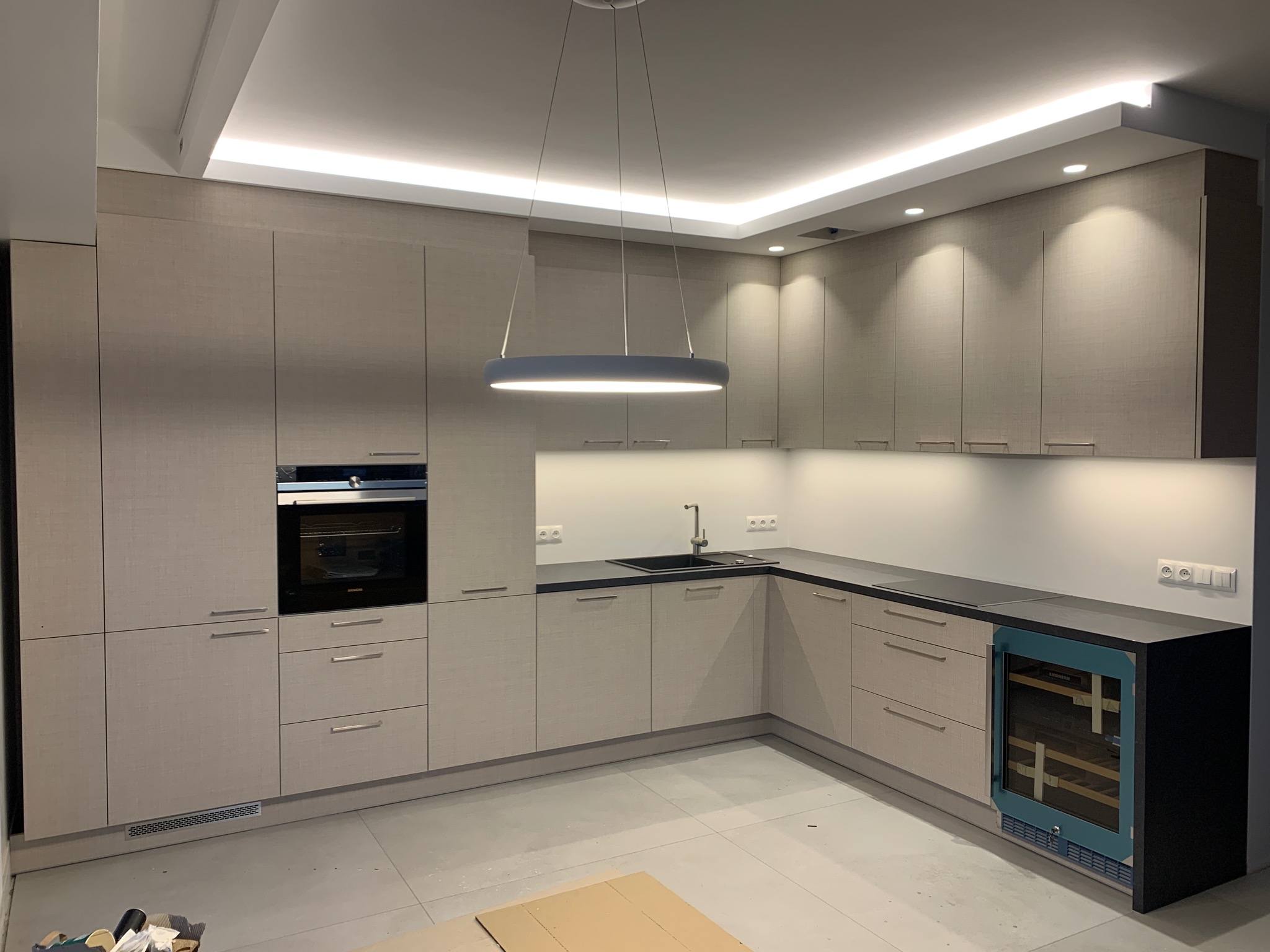 Nowoczesna kuchnia z jasnymi, teksturowanymi frontami szafek, czarnym blatem, okrągłą lampą wiszącą i podświetleniem LED podszafkowym oraz sufitowym, z wbudowanym piekarnikiem i chłodziarką do wina.