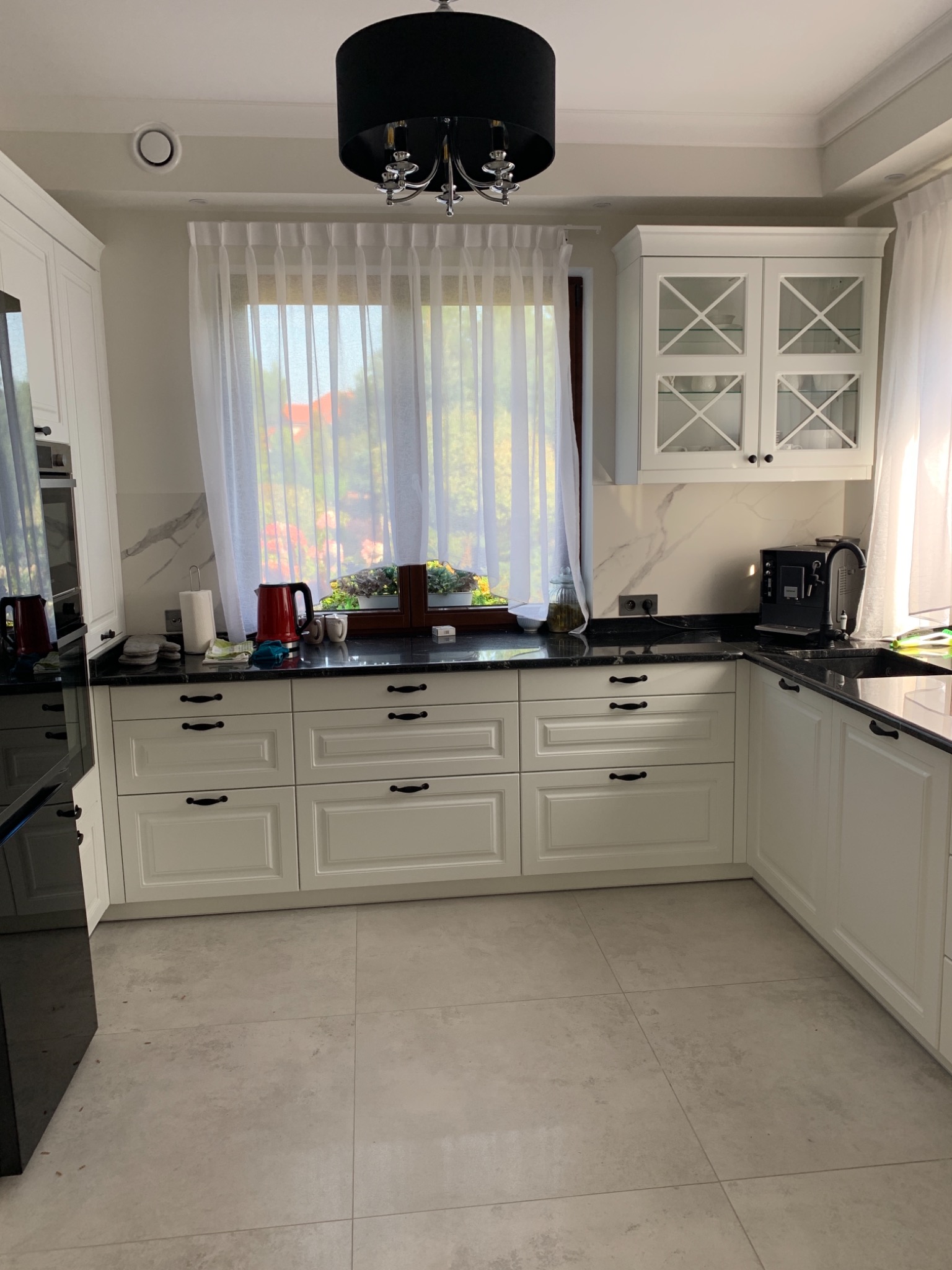 Jasna kuchnia z białymi szafkami, czarnym blatem i marmurowym wykończeniem ściany, okno z firanką i kwiatami, nowoczesna czarna lampa wisząca.