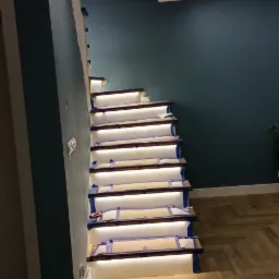 Montaż podświetlanych schodów drewnianych z zabezpieczeniem taśmą malarską, widoczne oświetlenie sufitowe i parkiet w jodełkę.