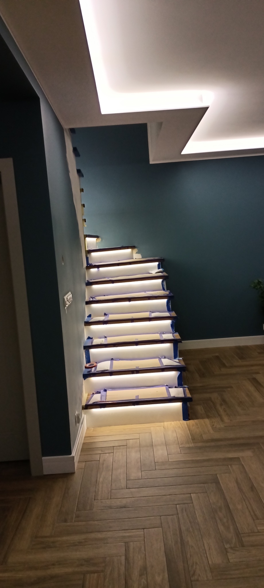 Montaż podświetlanych schodów drewnianych z zabezpieczeniem taśmą malarską, widoczne oświetlenie sufitowe i parkiet w jodełkę.