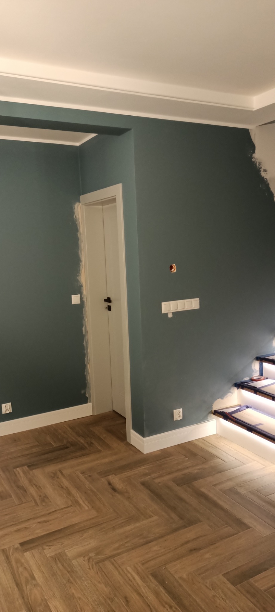 Pokój w trakcie malowania na kolor morski, widoczne ślady po taśmie malarskiej przy drzwiach i schodach, parkiet ułożony w jodełkę.