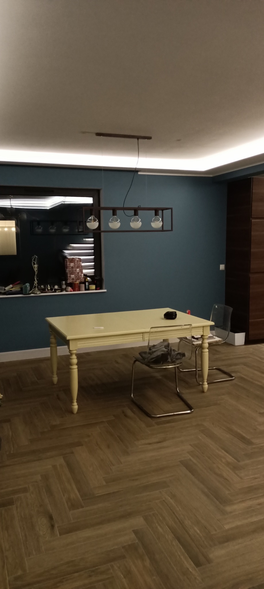 Jasny stół z toczonymi nogami i przezroczystymi krzesłami w pokoju z parkietem ułożonym w jodełkę i niebieskimi ścianami, oświetlonym designerską lampą wiszącą