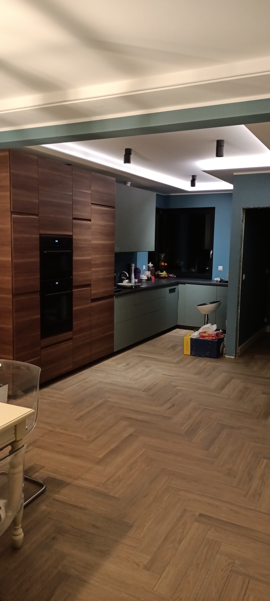 Nowoczesna kuchnia z drewnianymi szafkami, szarozielonymi frontami, parkietem w jodełkę i oświetleniem punktowym, w trakcie prac wykończeniowych, widoczny karton z narzędziami.