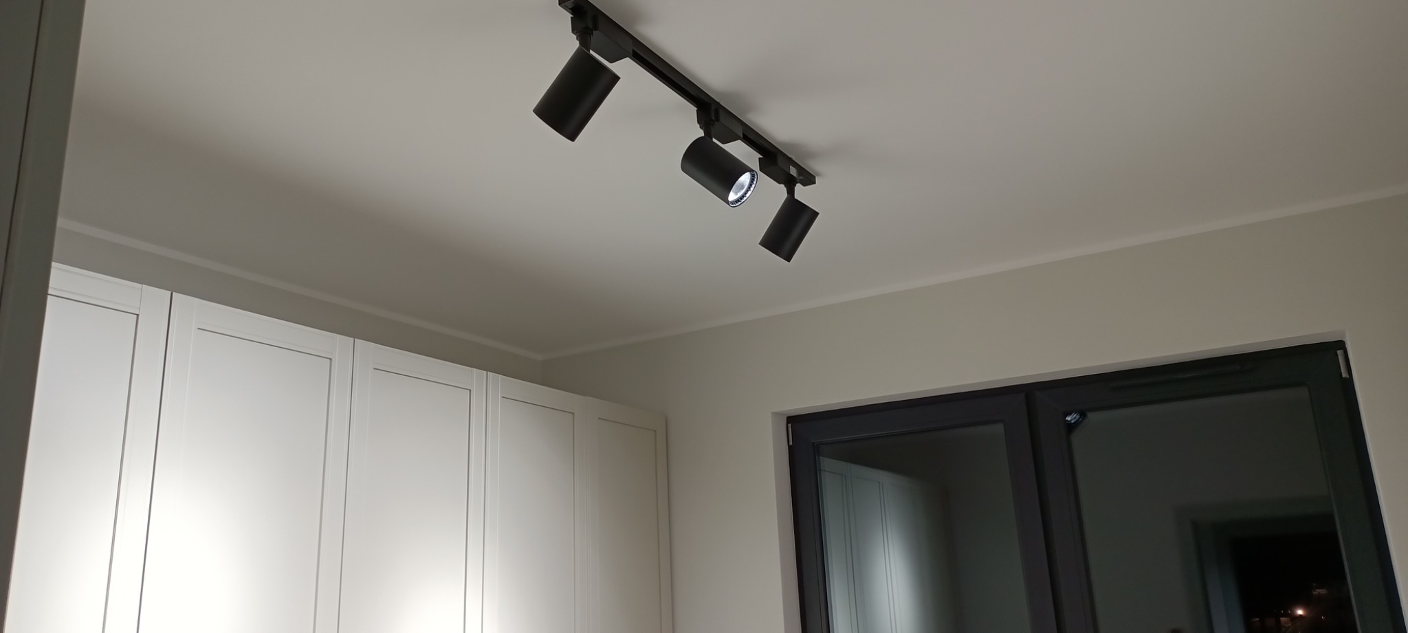 Nowoczesne oświetlenie sufitowe z trzema czarnymi reflektorami na szynie, skierowanymi w różne strony, na tle białych szafek i szarego okna z widokiem na noc.