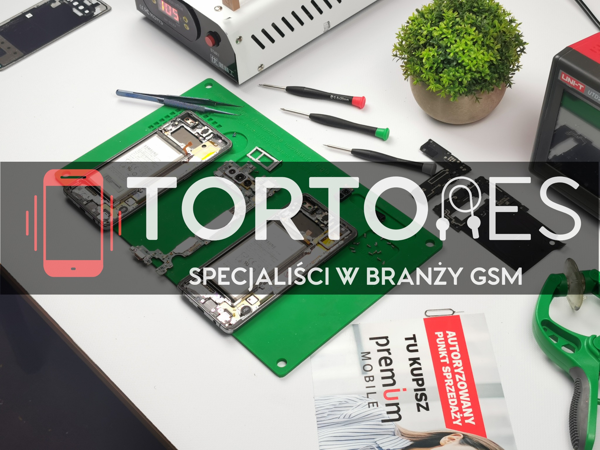 Rozłożone telefony na zielonej macie serwisowej, obok narzędzia serwisowe, penseta, śrubokręty, separator ekranu, roślina doniczkowa i ulotka reklamowa 'Premium Mobile'.