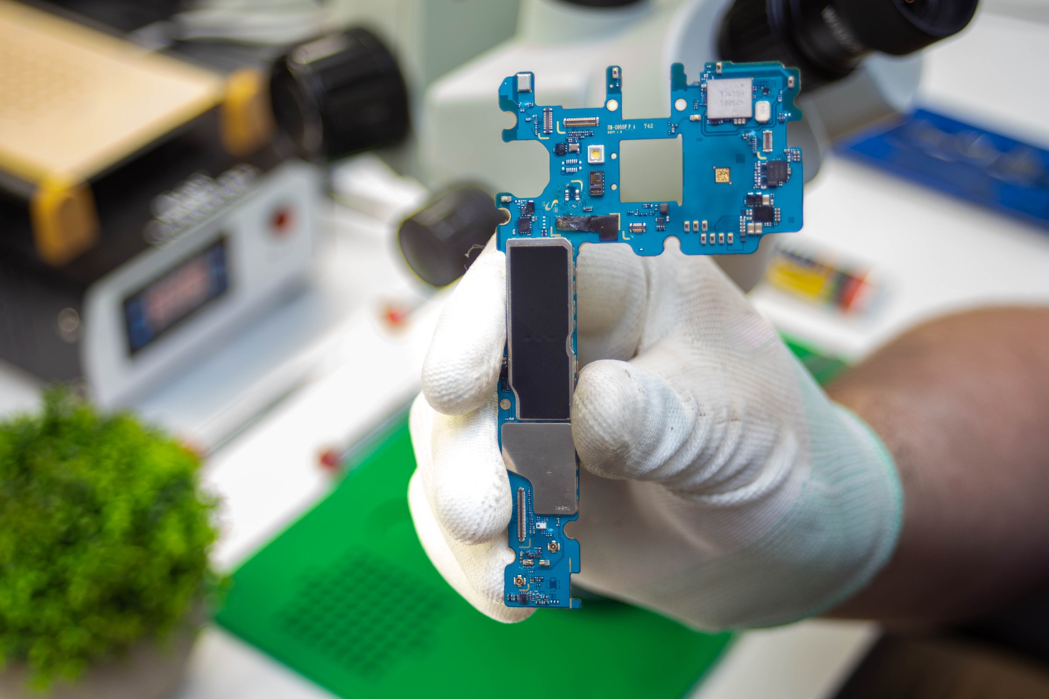 Płyta główna smartfona w białej rękawicy ochronnej, widoczny mikroskop i narzędzia serwisowe na stole.