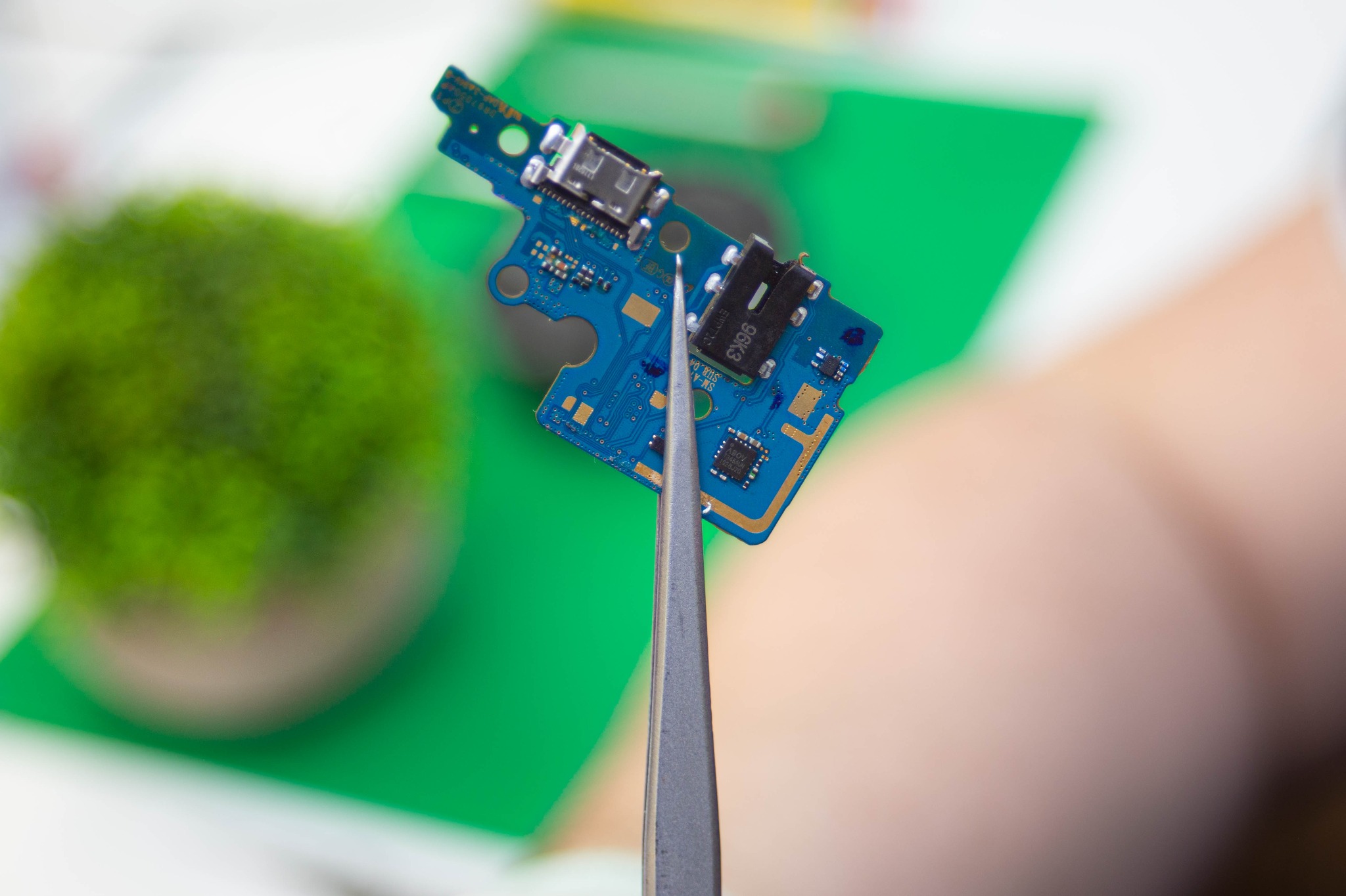 Niebieska płytka drukowana z portem ładowania USB-C, trzymana pincetą na rozmytym tle z elementami zieleni i beżu.