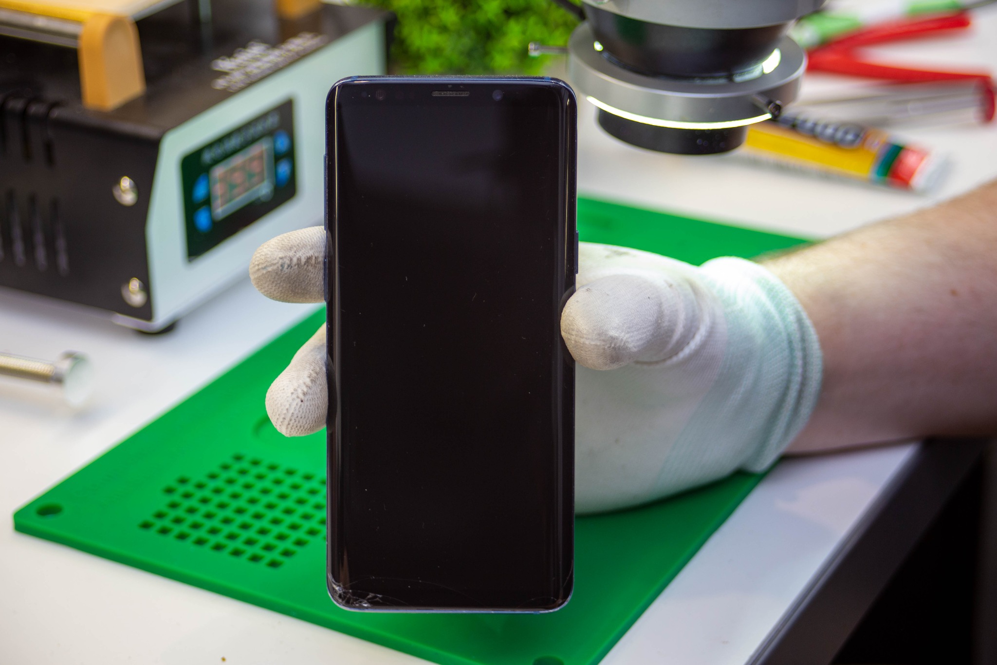 Uszkodzony smartfon trzymany w rękawicy ochronnej na zielonej macie, w tle sprzęt serwisowy i lupa.