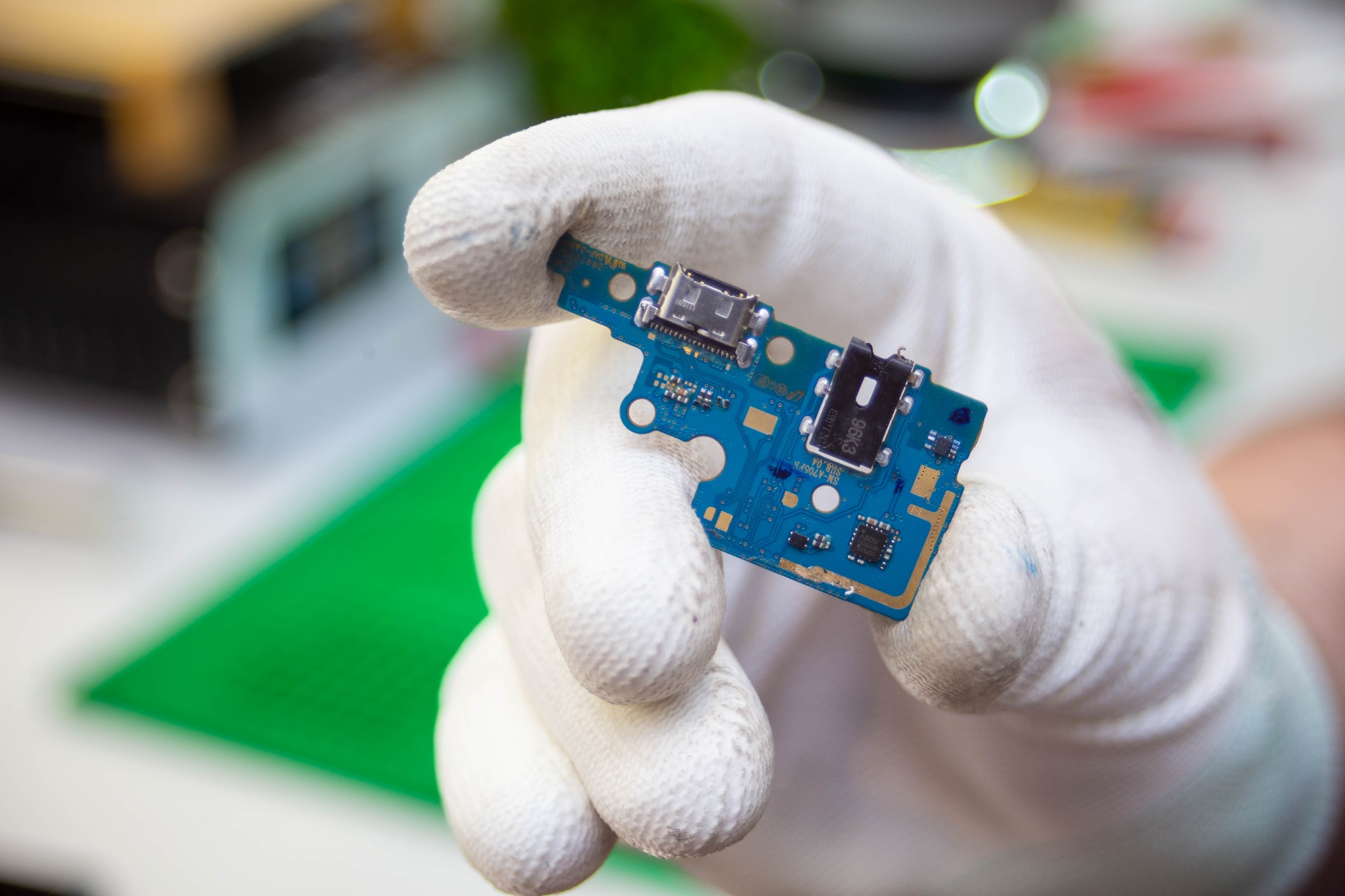 Błękitna płytka drukowana z portami USB trzymana w białej rękawicy ochronnej na rozmytym tle warsztatowym.