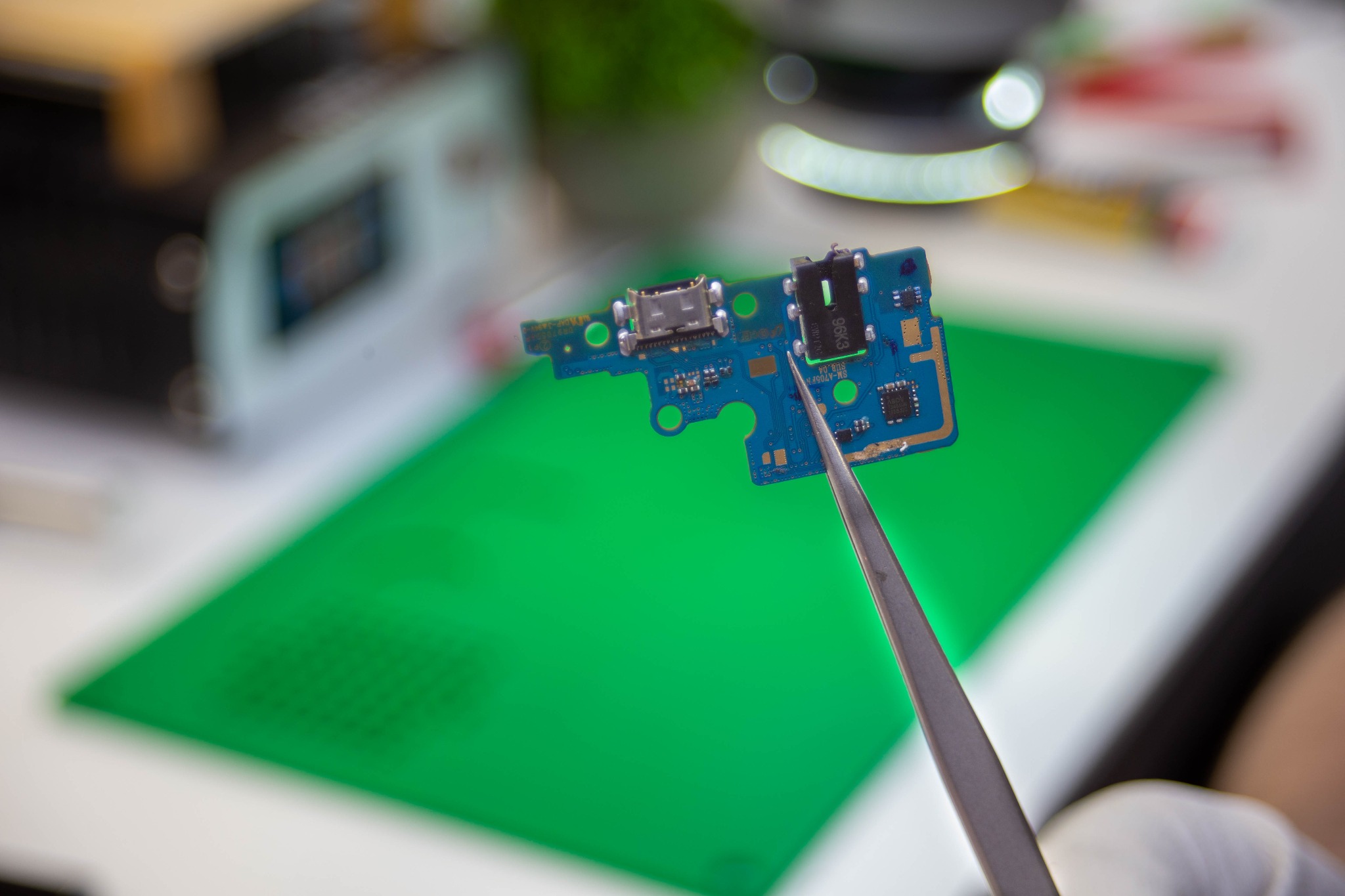 Płyta główna smartfona, trzymana pęsetą, z widocznymi portami USB-C i audio, na zielonej macie antyelektrostatycznej, w tle sprzęt serwisowy.