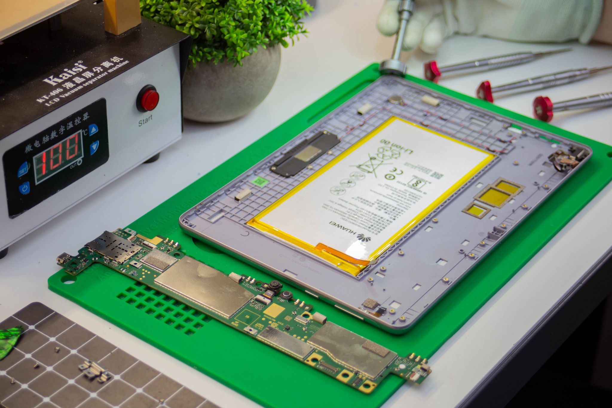Rozłożony tablet Huawei na zielonej macie serwisowej z widoczną baterią, płytą główną i narzędziami, obok maszyna do rozklejania ekranów.
